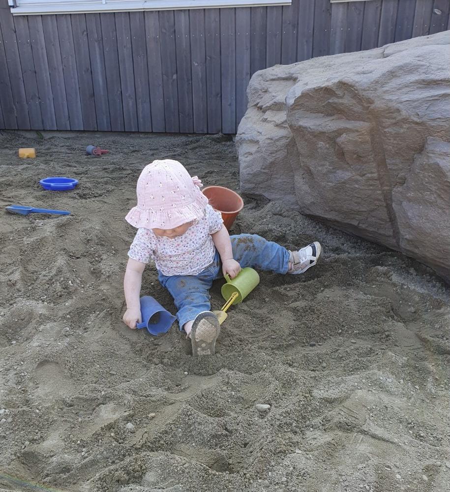 liten jente sitter i stor sanddunge og leker med spade og spann, har solhatt på